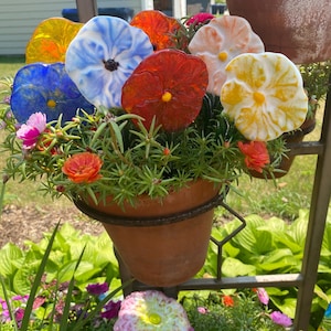 May include: A terracotta pot filled with green foliage and multiple fused glass flowers in vibrant colors including blue, yellow, red, and white. The glass flowers feature distinct centers and are arranged among small, natural orange and pink blossoms.
