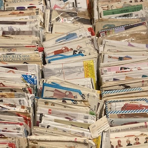 May include: A collection of vintage sewing patterns, featuring various brands and designs. The patterns are stacked together, showcasing a variety of illustrations and text, with prices visible on some of the pattern covers. The patterns are in various sizes and colors.