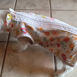 May include: A white dog vest featuring a colorful pattern of orange butterflies, pink flowers, yellow suns, and blue snails. The garment includes a full-length white zipper closure along the top, arm openings, and a high collar design for coverage.