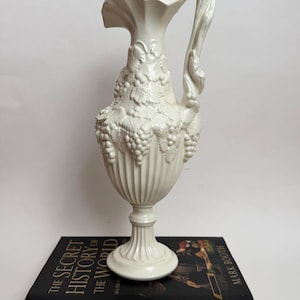 Może przedstawiać: Kremowy ceramiczny dzban z wysokim, żłobkowanym korpusem i zakrzywionym uchwytem. Dzban jest ozdobiony motywami winogron i liści. Stoi na książce zatytułowanej "The Secret History of the World".