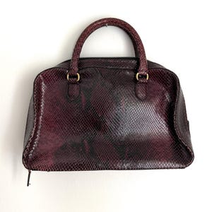 May include: A dark burgundy handbag featuring a textured snakeskin pattern across the exterior. The bag has two rounded top handles attached with gold-toned metal hardware and a zippered closure along the top edge of the structured frame.