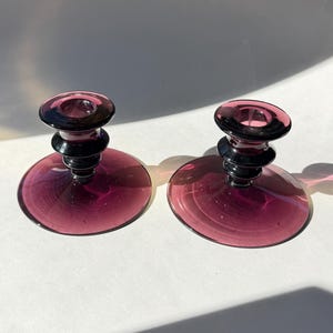 Może przedstawiać: Dwa pasujące fioletowe szklane świeczniki. Mają okrągłą podstawę i stopniowany trzon z okrągłą górą. Świeczniki wykonane są z przezroczystego fioletowego szkła z czarnym trzonem. Średnica podstawy ok. 10 cm.