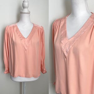 Puede incluir: Una blusa color melocotón con cuello en V y mangas largas. El escote y los detalles de los hombros tienen un acabado satinado. La blusa se exhibe en un maniquí blanco.