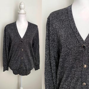 May include: A gray cardigan sweater with a V-neck and long sleeves. The sweater has a textured appearance and is fastened with four round, silver-colored buttons. The garment is displayed on a white mannequin.