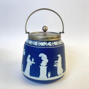 Op de afbeelding: Een blauwe en witte keramische koekjespot met een zilveren deksel en handvat. De pot heeft een wit cameo-ontwerp met figuren en een bloemmotief.