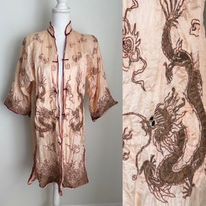 May include: A peach-colored, long-sleeved robe with a mandarin collar and dark brown dragon embroidery. The robe features a button closure and dark brown trim along the edges. The dragon design is visible on the front and sleeves.