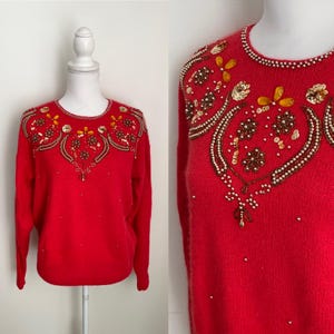 Puede incluir: Un suéter rojo brillante con intrincados adornos de cuentas y lentejuelas alrededor del cuello y los hombros. El diseño presenta patrones florales y abstractos en oro, marrón y blanco. El suéter se exhibe en un maniquí blanco.