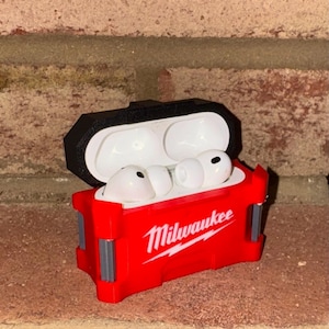 May include: Red and black protective case for wireless earbuds, with the Milwaukee logo in white script. The case is open, showing white earbuds. The case is made of a durable material.
