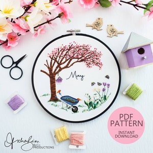 以下が含まれることがあります： 花が咲いた木、手押し車、花をモチーフにした刺繍のフープアート。フープは白で、外側のリングは黒。フープの中央には「5月」の文字が刺繍されている。