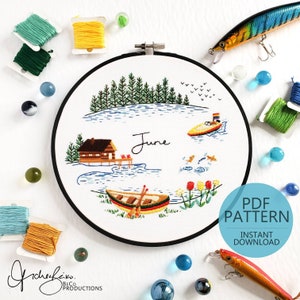 May include: Embroidered hoop art featuring a lake scene with a cabin, a boat, and the word "June". The hoop is surrounded by colorful embroidery floss and fishing lures.