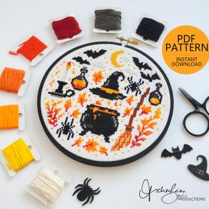 Pode incluir: Um padrão de ponto cruz com tema de Halloween em um bastidor redondo, com um chapéu de bruxa, caldeirão, morcegos e folhas de outono. A imagem inclui carretéis de linha de bordar laranja, vermelha, amarela e cinza, junto com tesouras e o texto "PDF Pattern - Instant Download."