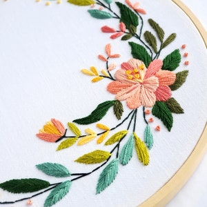 May include: A floral embroidery hoop art piece with a wreath of green, yellow, and pink flowers. The embroidery is on white fabric and is framed in a natural wood hoop.
