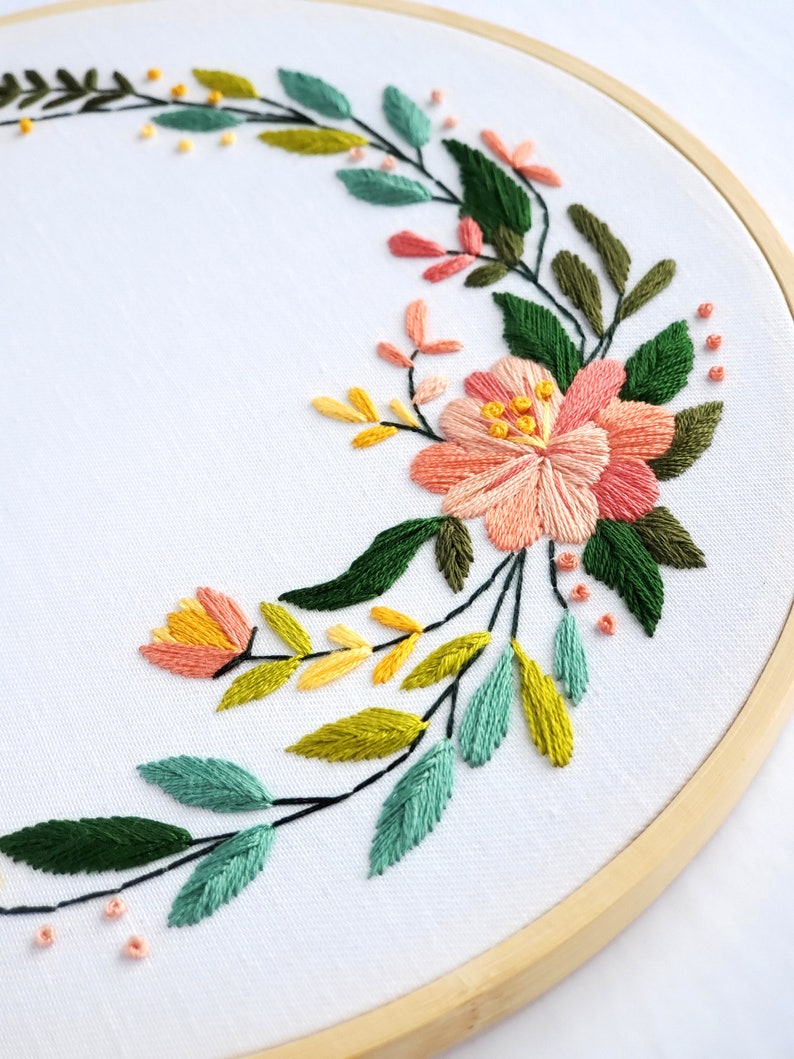 May include: A floral embroidery hoop art piece with a wreath of green, yellow, and pink flowers. The embroidery is on white fabric and is framed in a natural wood hoop.