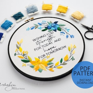 May include: A white embroidery hoop with a black frame, featuring a floral design with yellow, blue, and green embroidery floss. The text "Wishing you strength for today and hope for tomorrow" is embroidered in black thread.