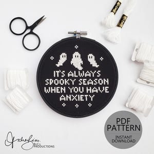 May include: A black cross-stitch hoop with the text "It's Always Spooky Season When You Have Anxiety" and three ghost figures. White thread, scissors, and a PDF pattern graphic are also visible.