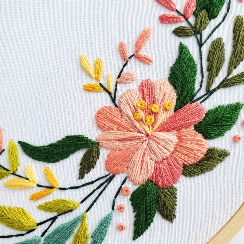 May include: Embroidered floral hoop art with a pink flower and green leaves. The flower has a yellow center with a delicate stitch pattern.