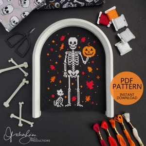 May include: A cross-stitch pattern featuring a skeleton holding a pumpkin, with a cat, and autumn leaves on a black background. The design is framed in a white arch. Spools of thread and bone decorations are also visible.