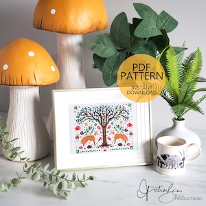 May include: A framed cross-stitch pattern featuring a tree with two foxes, mushrooms, and floral elements. The image is in a gold frame. The words "PDF PATTERN INSTANT DOWNLOAD" are in a circular graphic. Decorative ceramic mushrooms and a mug are also in the image.