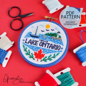 Puede incluir: Un proyecto de punto de cruz en un bastidor azul con un velero, un faro y una hoja de arce roja con el texto FOREVER LAKE ONTARIO. El diseño está rodeado por unas tijeras negras y varios carretes de hilo de bordar de colores.