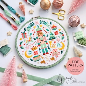 Puede incluir: Un bastidor de bordado con un diseño navideño que incluye un cascanueces, un árbol de Navidad y otros elementos festivos. El diseño está rodeado de hilo verde y otros suministros de manualidades, incluyendo adornos rosas y dorados, e hilo de bordar.