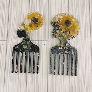 以下が含まれることがあります： 金色のグリッター仕上げの黒いヘアコーム2本。各コームは、押し花の花冠をかぶった黒人女性の顔のレジンデザインが施されています。1つのコームは黄色と白の花冠、もう1つは黄色の花冠です。