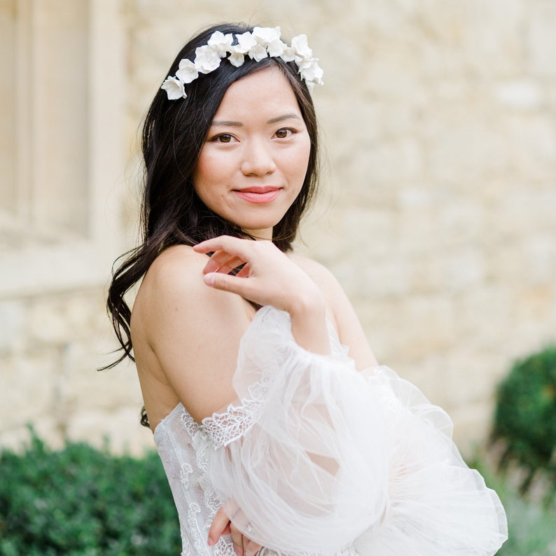 May include: A woman wearing a white off-the-shoulder dress with puffy sleeves and a flower crown. She is standing in front of a stone wall.