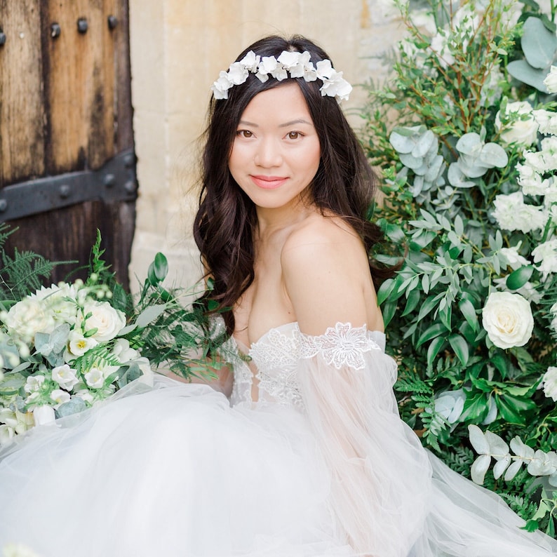 May include: A bride wearing a white off-the-shoulder gown with lace detailing and a flower crown. She is sitting in front of a wall of greenery and white flowers.