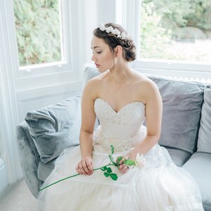 May include: A bride wearing a white strapless wedding dress with a floral crown and holding a single white rose.