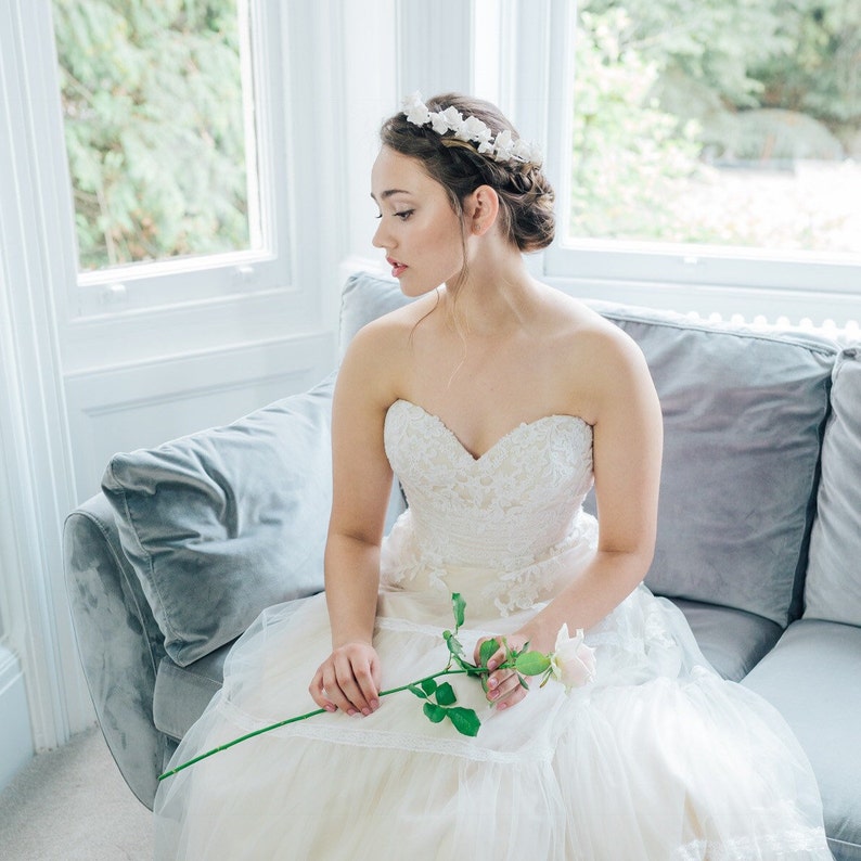 May include: A bride wearing a white strapless wedding dress with a floral crown and holding a single white rose.