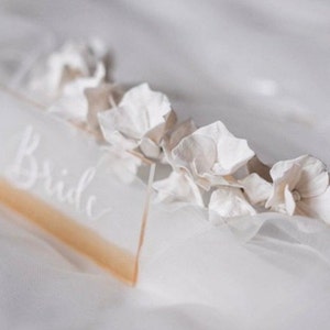 May include: A white acrylic sign with the word "Bride" written in calligraphy. The sign is decorated with white flowers.