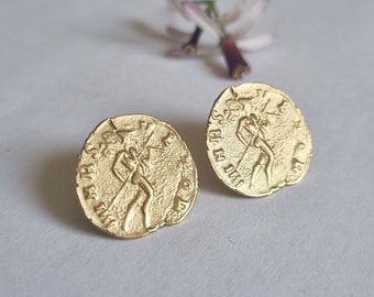 May include: A pair of gold-toned earrings featuring a circular design with a raised image of a person holding a spear. The earrings are approximately 1 inch in diameter.