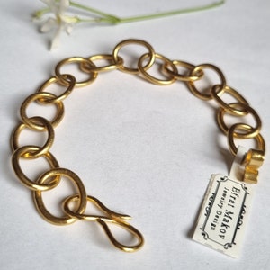 Puede incluir: Un brazalete de cadena dorada con cierre. El brazalete está hecho de eslabones redondos y gruesos. La etiqueta adjunta al brazalete dice "Efrat Makov Jewelry Design".