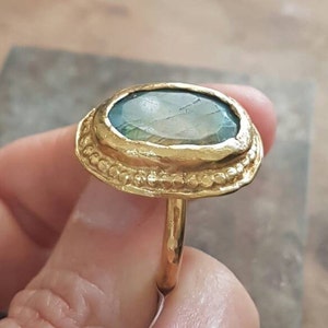May include: A gold ring with a large oval green gemstone set in a textured gold bezel. The ring has a beaded edge and a simple band.