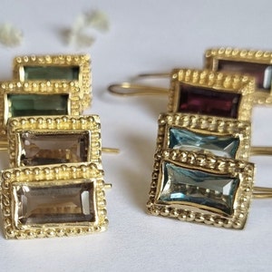 Puede incluir: Un conjunto de pendientes de oro con piedras preciosas de diferentes colores. Los pendientes son de forma rectangular y tienen un borde de cuentas. Las piedras preciosas son verdes, marrones, moradas y azules.