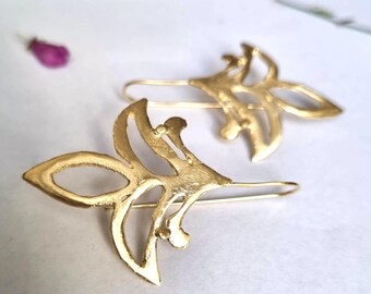 May include: A pair of gold-toned earrings with a delicate, intricate design. The earrings are shaped like stylized leaves with cut-out details.