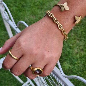 Puede incluir: Un brazalete de cadena dorada con un dise&ntilde;o de hoja de oro. Un anillo de oro con una piedra preciosa marr&oacute;n est&aacute; en el dedo &iacute;ndice.