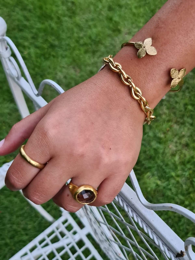 Puede incluir: Un brazalete de cadena dorada con un dise&ntilde;o de hoja de oro. Un anillo de oro con una piedra preciosa marr&oacute;n est&aacute; en el dedo &iacute;ndice.