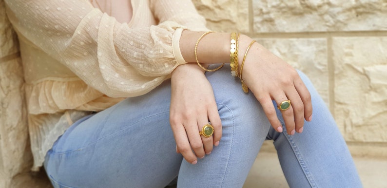 Puede incluir: Una persona lleva una blusa de gasa beige claro con un brazalete de cadena dorada y dos anillos de oro con piedras verdes.