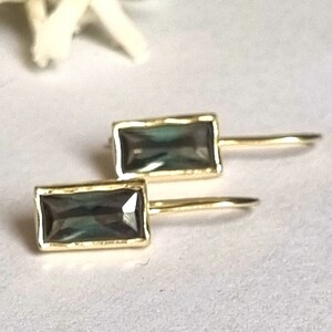 May include: A pair of gold earrings with rectangular, dark grey gemstones. The earrings have a simple, minimalist design.