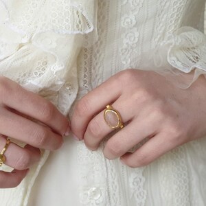 Puede incluir: Una mano de mujer con un anillo de oro con una piedra ovalada de color rosa claro. El anillo est&aacute; en el dedo medio de la mano. La mano lleva un vestido blanco de encaje.