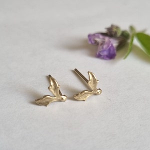 May include: A pair of gold-toned stud earrings shaped like small, textured leaves. Each earring features a delicate, organic form with a pointed tip and a post backing, designed to sit flush against the earlobe when worn as a set of two.