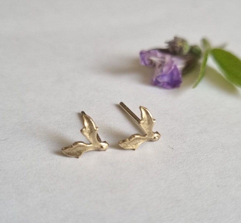 May include: A pair of gold-toned stud earrings shaped like small, textured leaves. Each earring features a delicate, organic form with a pointed tip and a post backing, designed to sit flush against the earlobe when worn as a set of two.