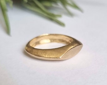 May include: A gold ring with a flat, rectangular top and rounded edges. The ring is made of a smooth, brushed metal.