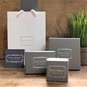 May include: A set of five gray boxes with the text "Efrat Makov Jewelry Design" printed on each box. The boxes are arranged in a pyramid shape, with the largest box at the bottom and the smallest box at the top. The boxes are on a wooden surface.