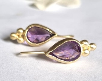 May include: A pair of gold earrings with teardrop-shaped amethyst stones. The earrings have a delicate design with small gold beads.