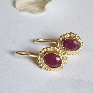 Puede incluir: Un par de pendientes de oro con piedras preciosas de rubí. Los pendientes tienen un diseño delicado y con cuentas alrededor de las piedras de forma ovalada.