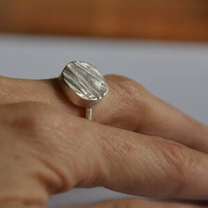 May include: A silver ring with a textured oval-shaped top. The ring is worn on a finger.