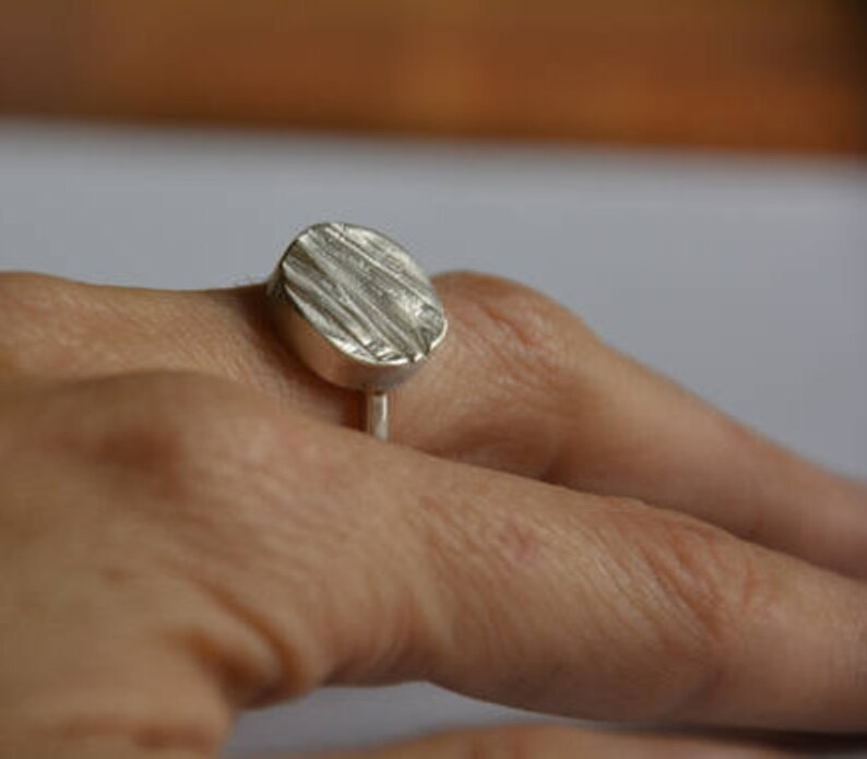 May include: A silver ring with a textured oval-shaped top. The ring is worn on a finger.