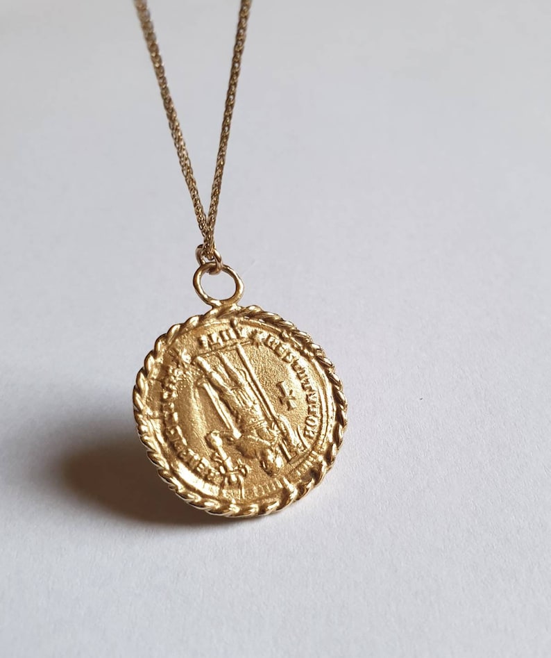 May include: Gold pendant necklace with a round, detailed medallion featuring a religious figure. The medallion has a rope-like border and inscription.