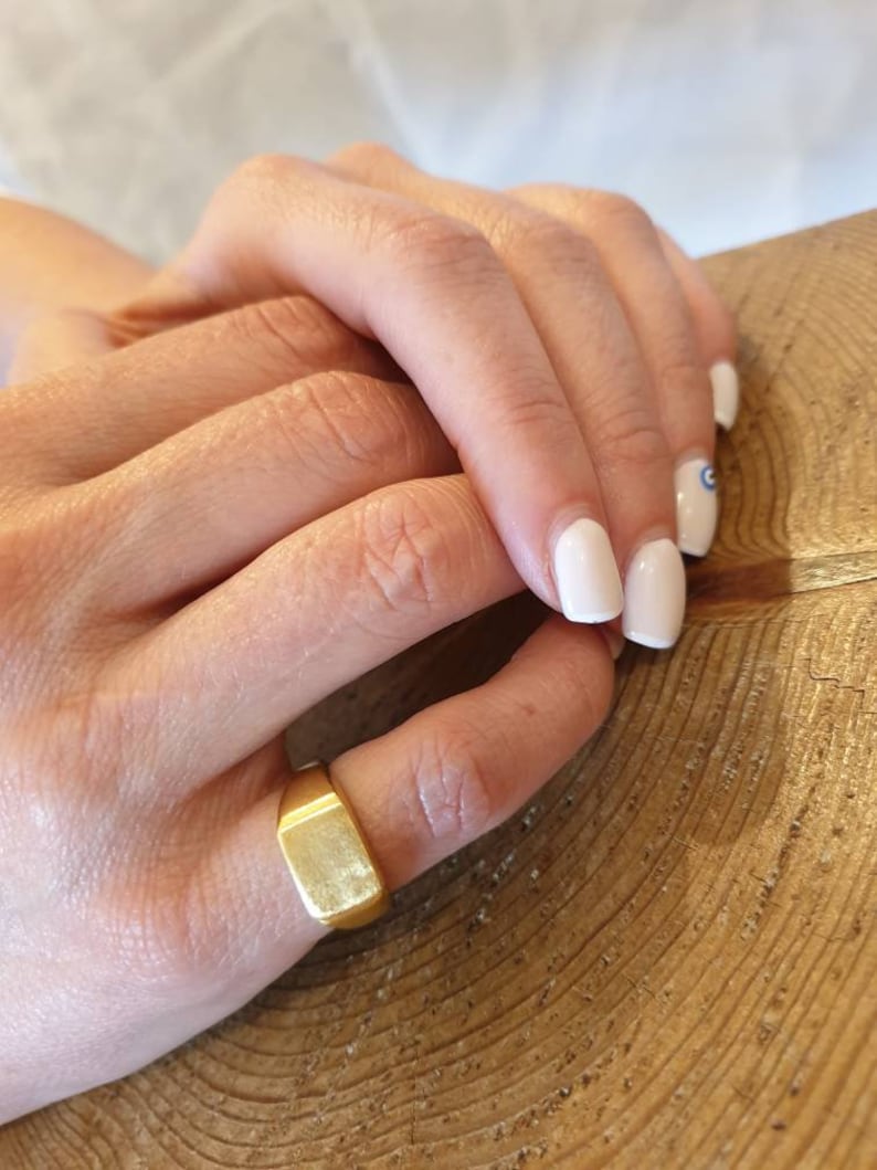 May include: A gold signet ring with a flat, rectangular top. The ring is worn on a finger.